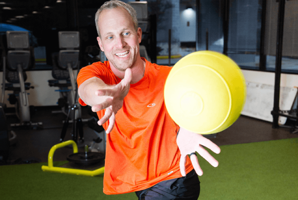 A man in an orange shirt smiles while preparing to catch a yellow ball in a strength training exercise for longevity