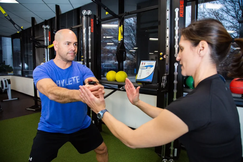 A man in a blue shirt completes an arm strength workout while being coached by a female trainer.