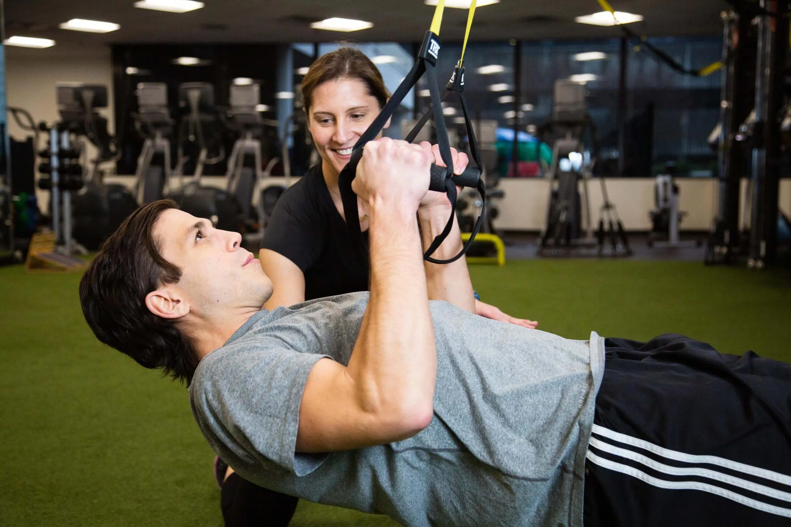 A female personal trainer catches a male client while he holds onto a TRX machine